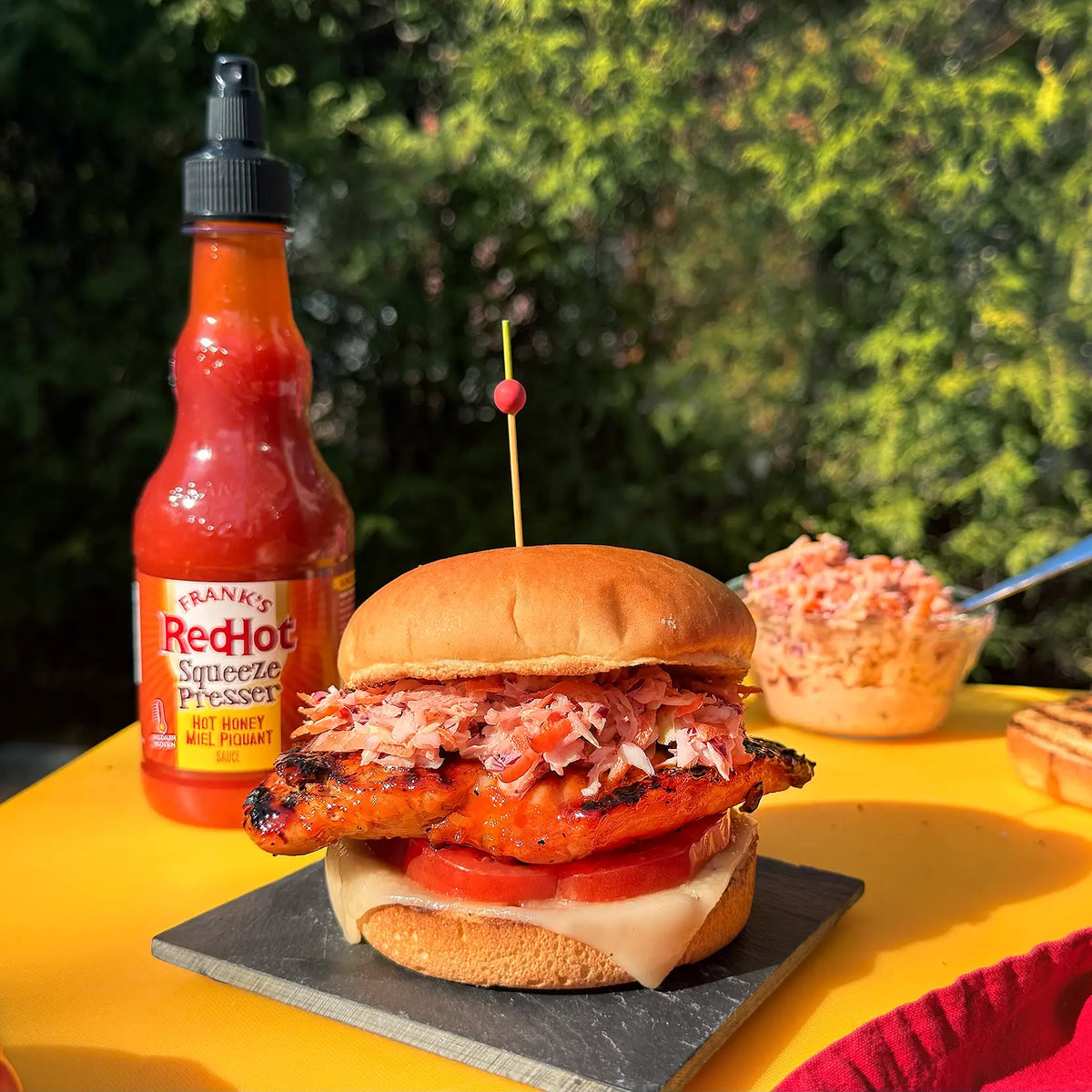 Frank’s RedHot Squeeze Hot Honey Sauce beside a grilled chicken sandwich topped with slaw
