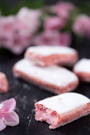 Pink rectangular cookies dusted with white powdered sugar, with one broken piece showing a pink crumb interior, set on a dark surface with blurred pink flowers in the background.