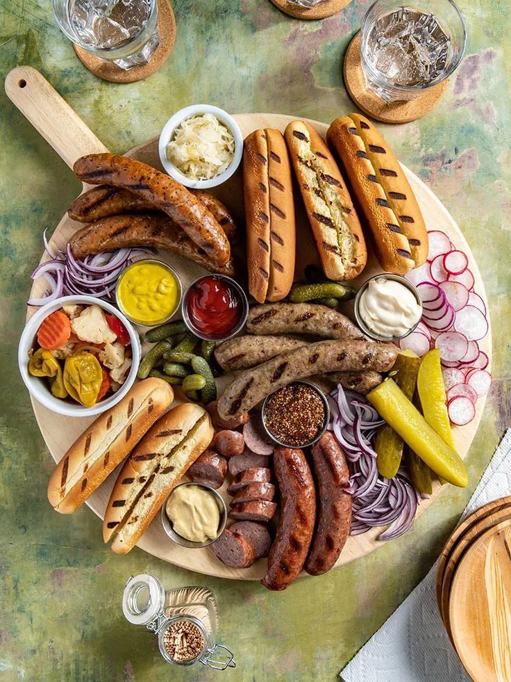Overhead view of a serving board with sausages, sausage sandwiches, pickles, sliced onions and condiments.