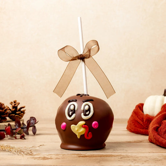 A novelty Halloween candy apple decorated to look like a turkey, featuring a brown chocolate body, white eyes, pink cheeks, a yellow beak, and a red wattle, complete with a white stick and a brown ribbon bow on top, set on a wooden surface with autumn props.