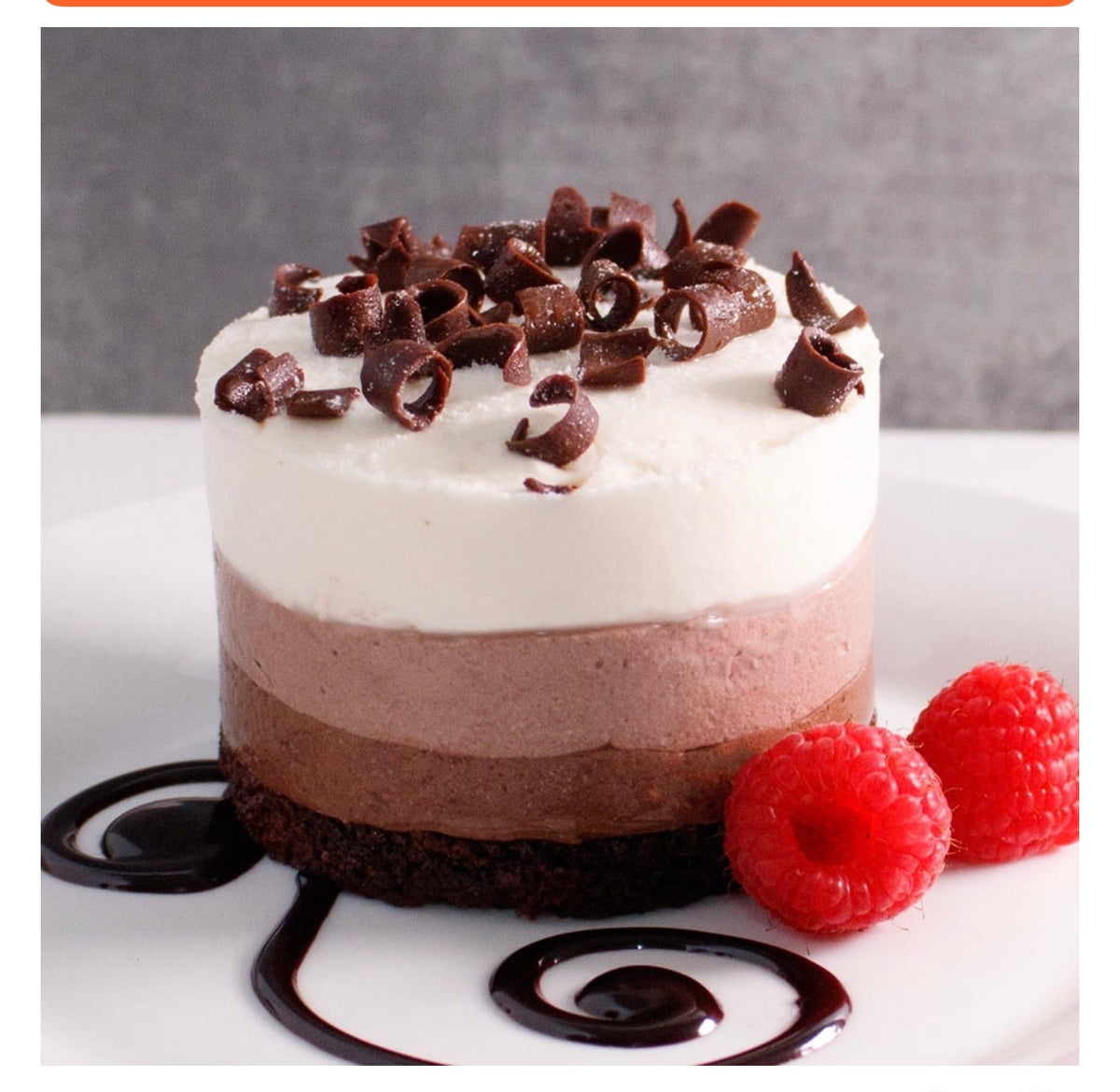 A small layered dessert cake with white cream, chocolate mousse, and a dark chocolate base, topped with chocolate curls, served on a white plate with raspberries and chocolate drizzle against a gray background.