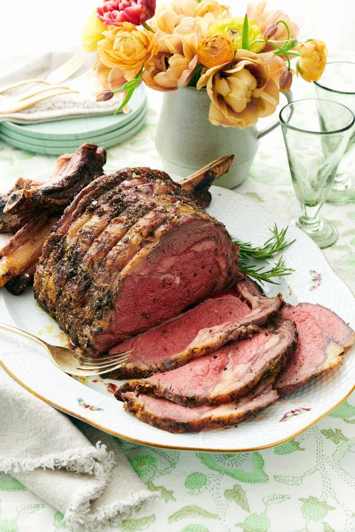 A sliced roasted beef or cooked prime rib roast on a white oval platter with a gold rim, set on a patterned tablecloth with a vase of yellow flowers, plates, and a glass in the background.