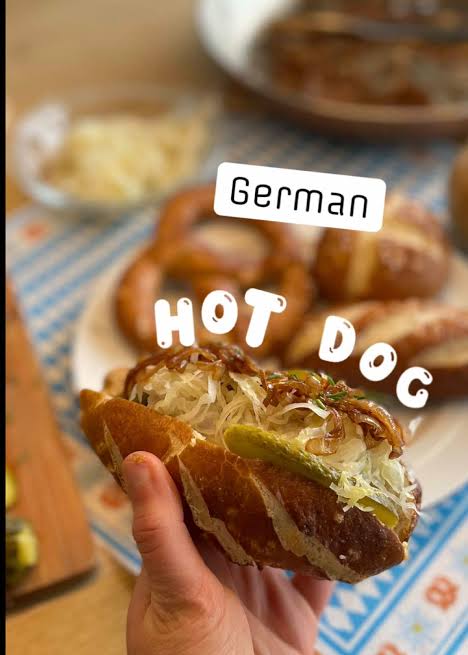 Hand holding a sausage-filled pretzel roll with cabbage beneath German Hot Dog text