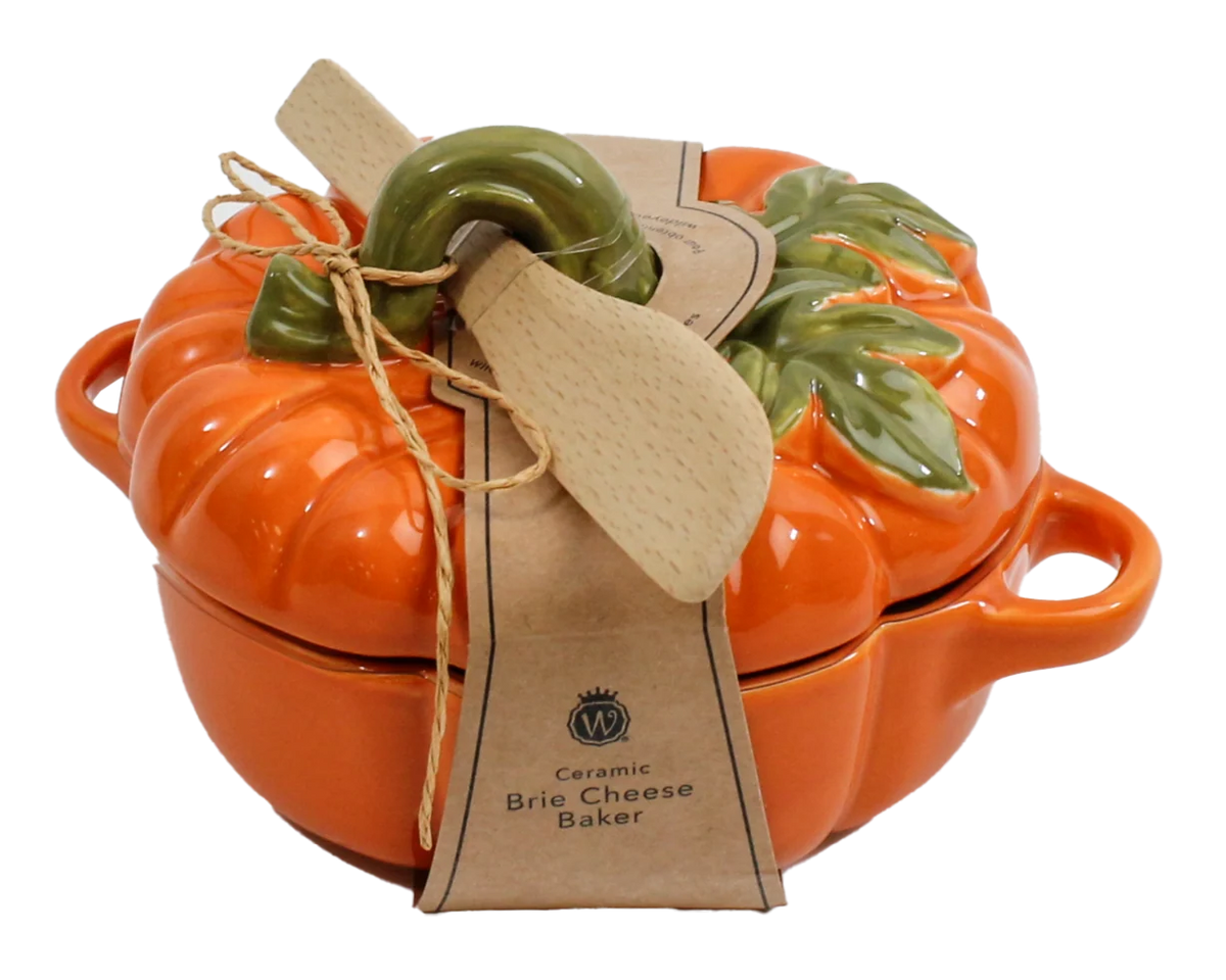 An orange ceramic pumpkin-shaped brioche cheese baker with a matching lid, decorated with a green ceramic stem and leaves. It is wrapped in a kraft paper ribbon with a wooden spoon resting on top, set against a plain white background.
