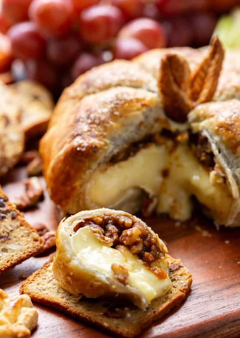 A close-up of a baked cheese stuffed pastry or bread roll on a wooden board, with a piece broken open to show melted cheese and walnut topping, served with crackers and red grapes in the background.
