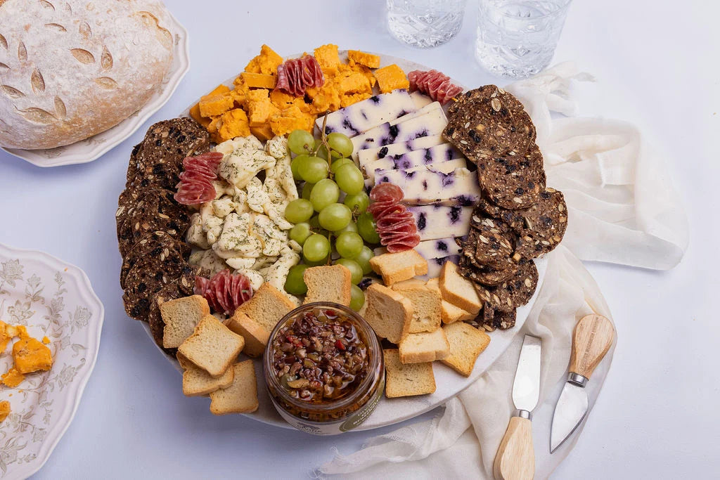Assorted cheese platter with crackers, fruits, and a loaf of bread on a white surface.