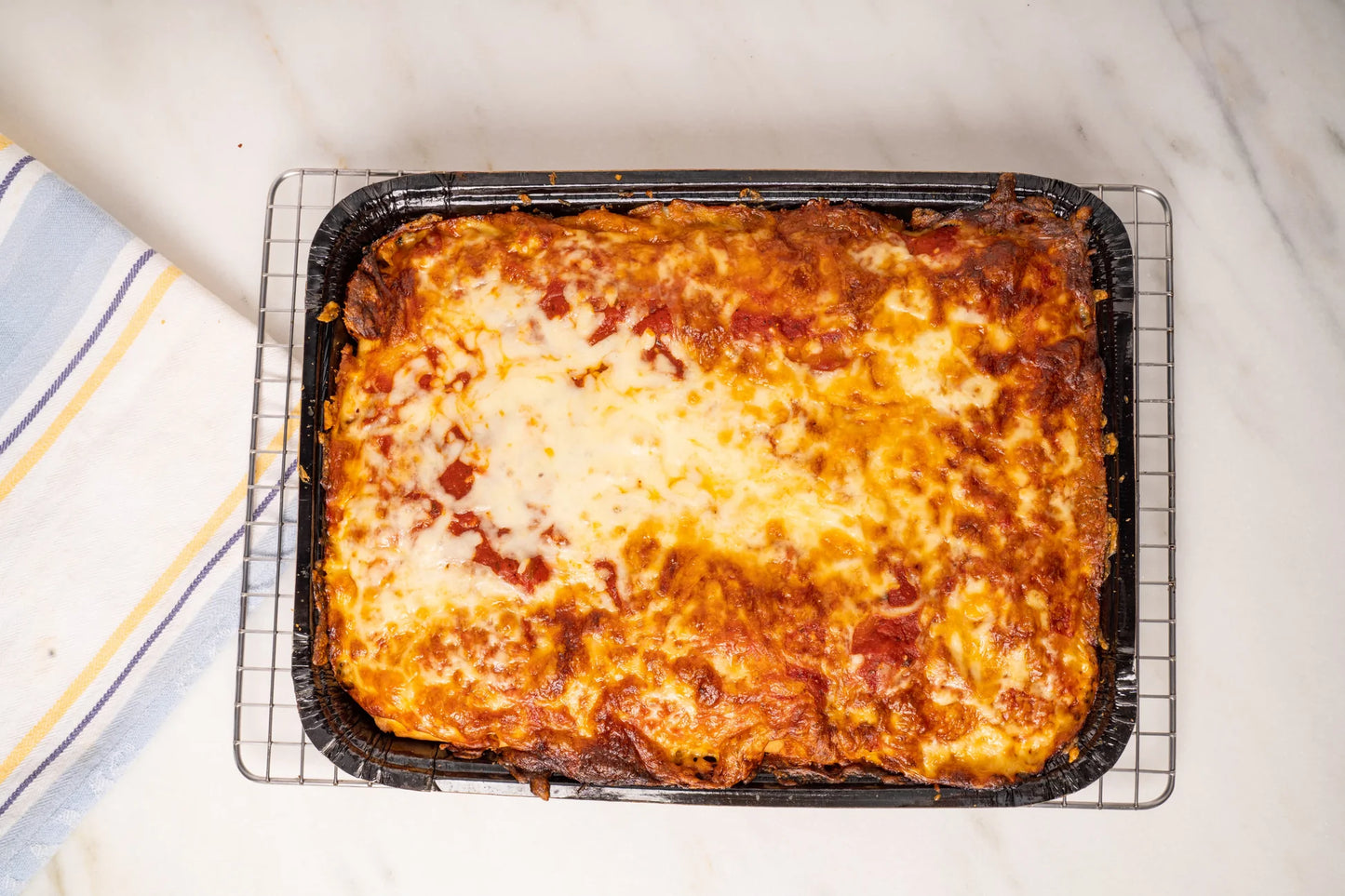 A baked rectangular lasagna or pasta dish in a black tray, resting on a wire cooling rack on a white marble surface with a striped kitchen towel partially visible at the left edge.