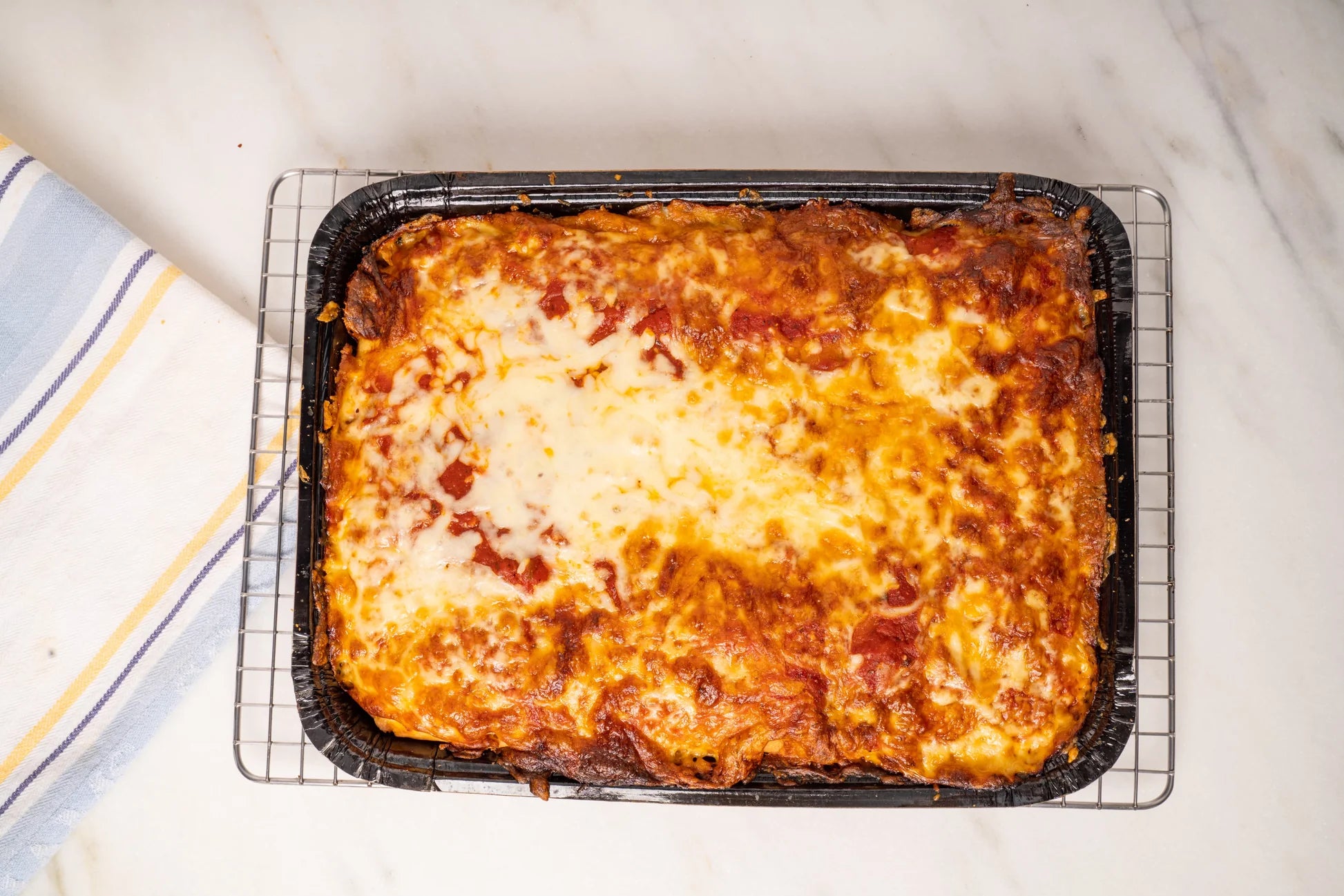 A baked rectangular lasagna or pasta dish in a black tray, resting on a wire cooling rack on a white marble surface with a striped kitchen towel partially visible at the left edge.