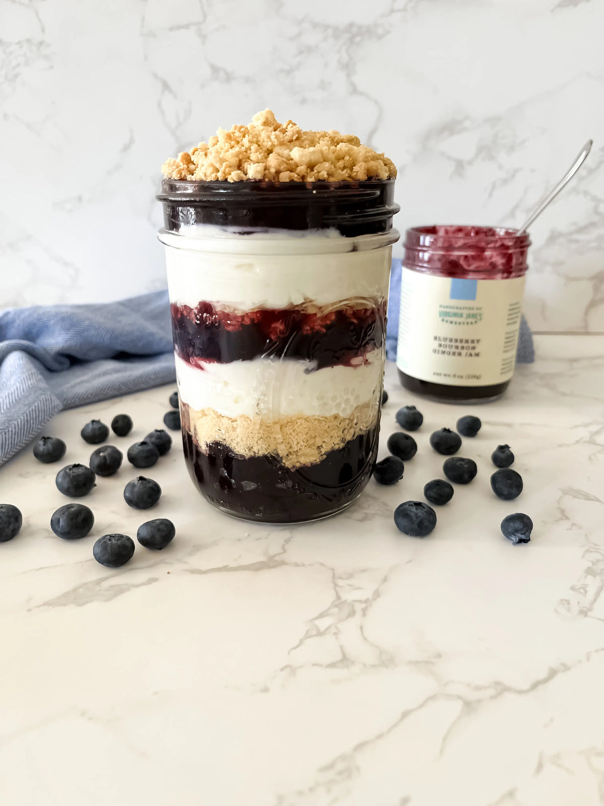 A layered blueberry and cream parfait in a clear glass mason jar with a crumb topping, styled on a marble surface with scattered blueberries and a jar of blueberry jam in the background.