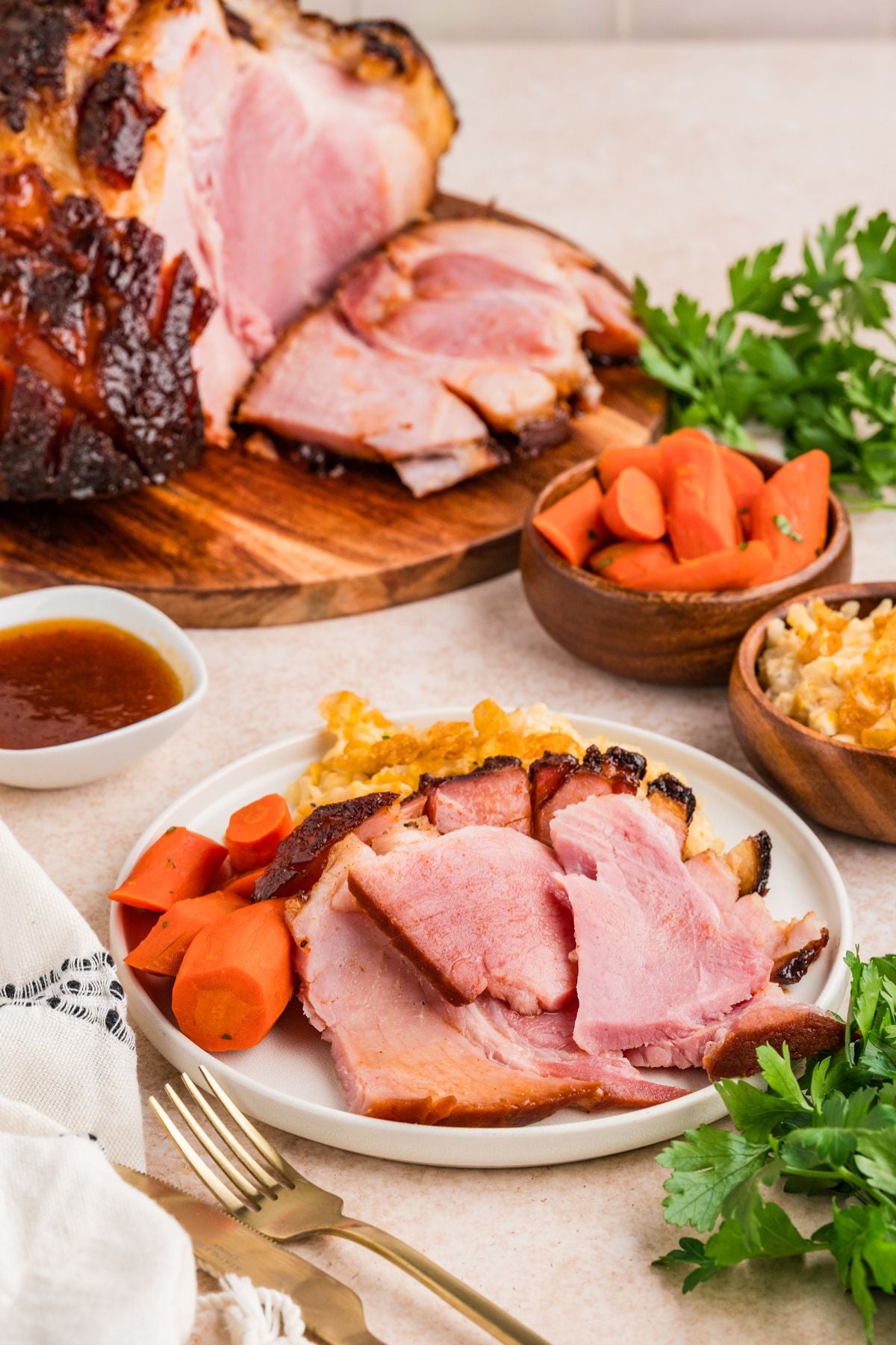 Sliced ham on a white plate with carrots, mashed potatoes, and a side of sauce, styled on a dining table with a whole ham, bowls of vegetables, and parsley.