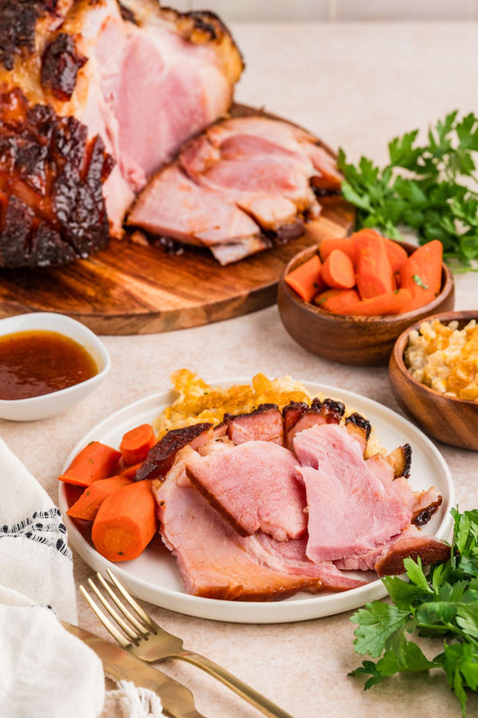 Sliced ham on a white plate with carrots, mashed potatoes, and a side of sauce, styled on a dining table with a whole ham, bowls of vegetables, and parsley.