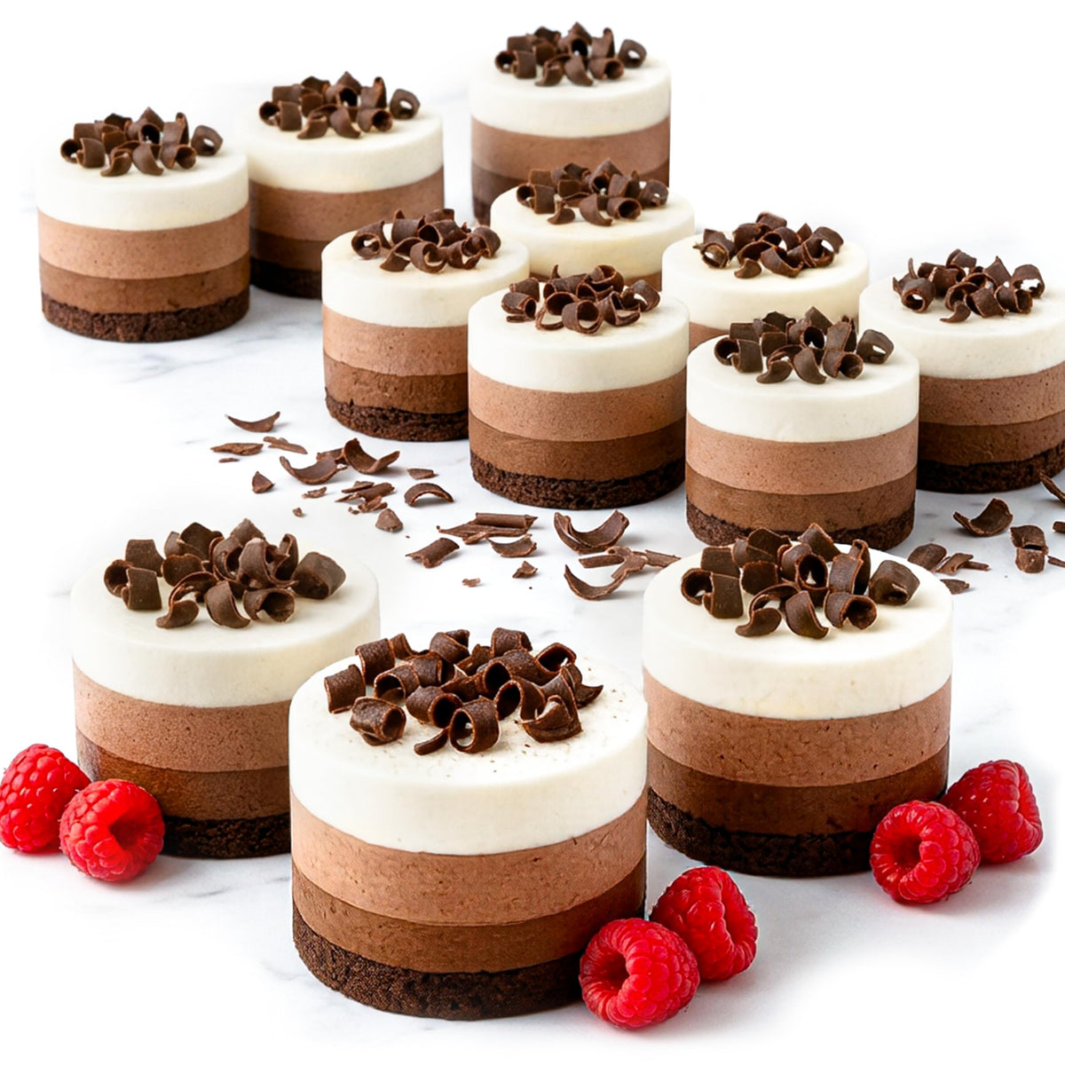Multiple mini layered dessert cakes topped with chocolate curls, arranged on a white marble surface with raspberries and scattered chocolate shavings.