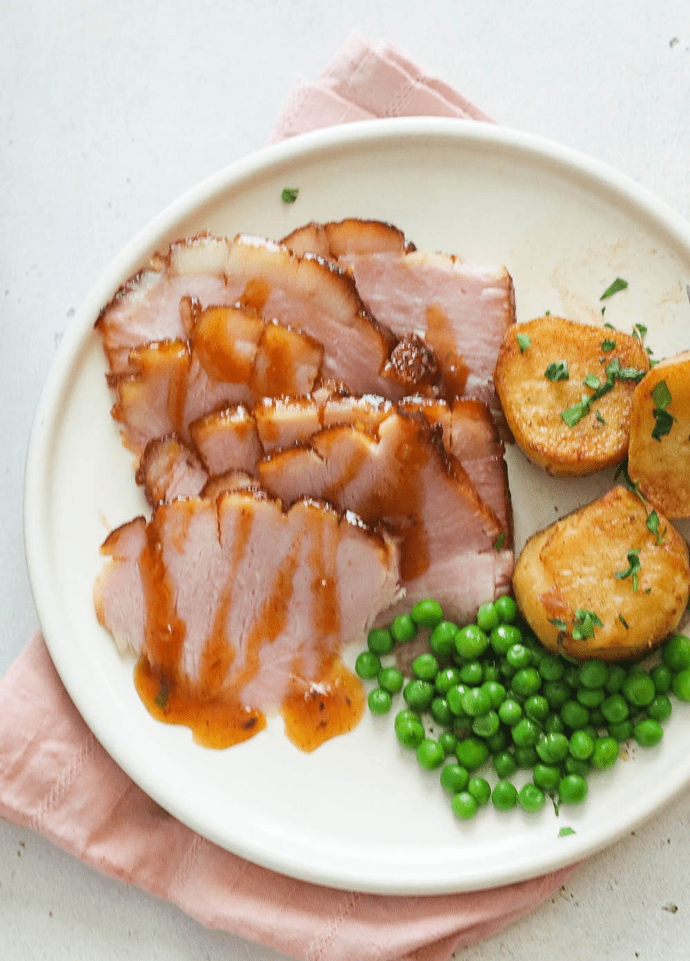 A top-down view of a plated meal featuring sliced glazed ham, green peas, and roasted potato wedges on a white plate with a pink napkin.