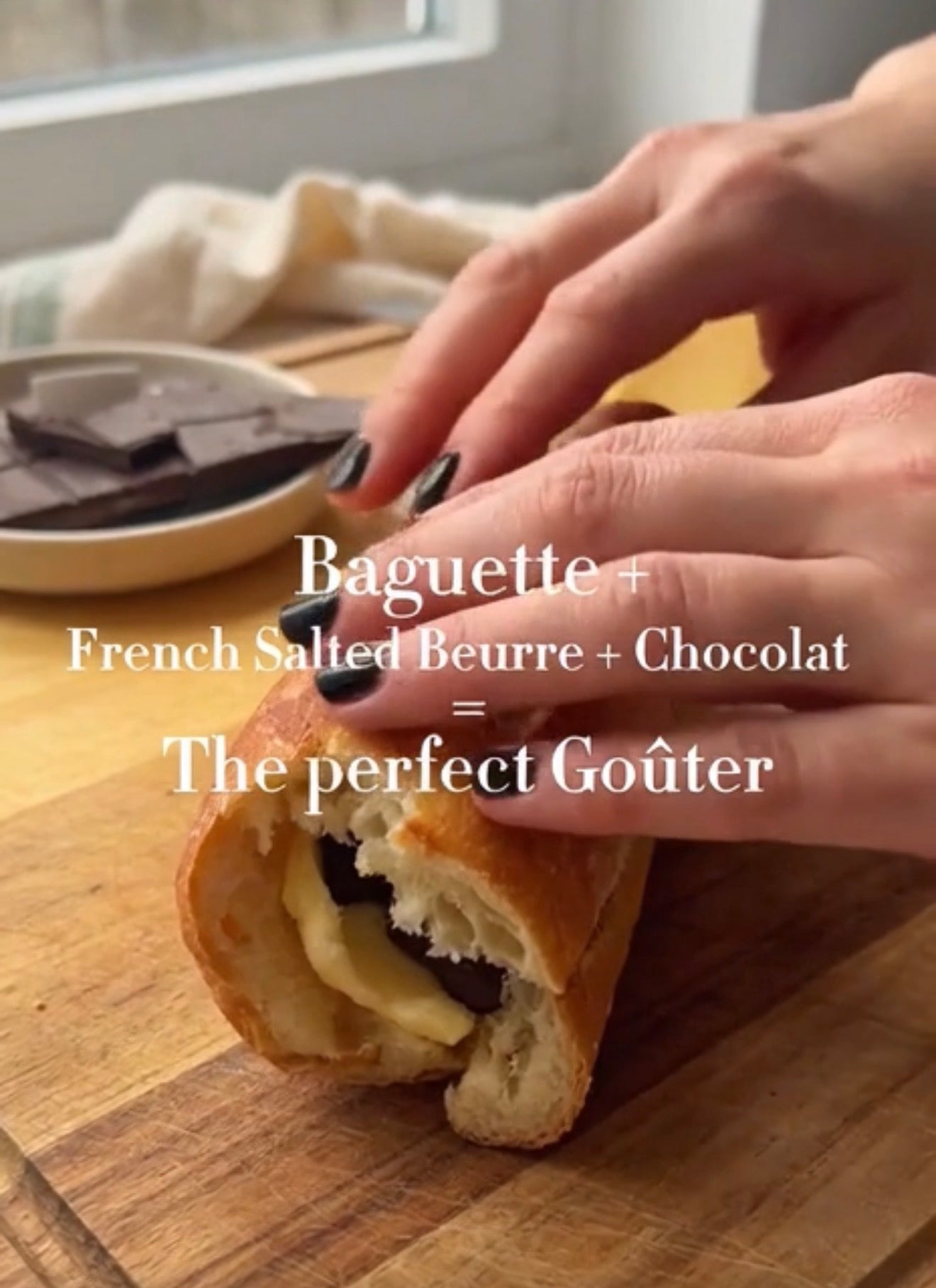 A close-up of a hand slicing a baguette filled with chocolate and cream on a wooden cutting board, with text overlay reading 'Baguette + French Salted Beurre + Chocolat = The perfect Goüter'.