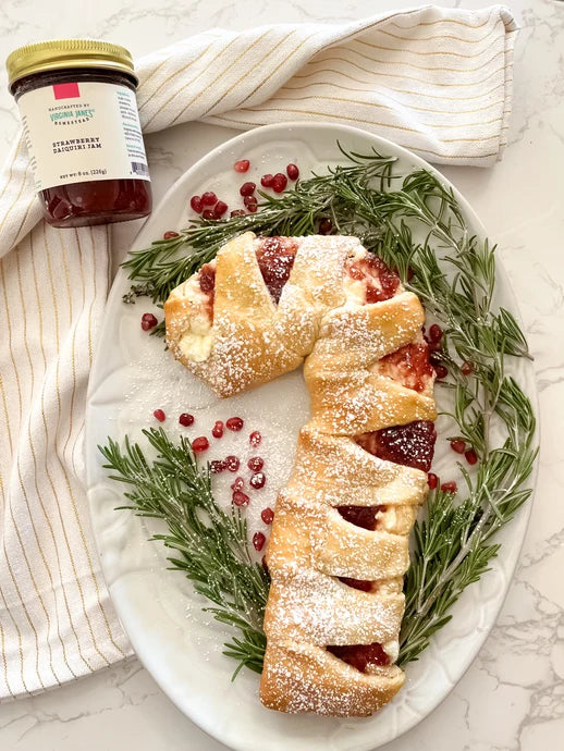 A festive holiday dessert, a braided strawberry cream cheese pastry, is presented on a white oval platter decorated with rosemary sprigs and pomegranate seeds. A jar of Strawberry Cream Cheese Jam by Sweet Home Jam is visible in the top left corner on a striped cloth over a marble surface.