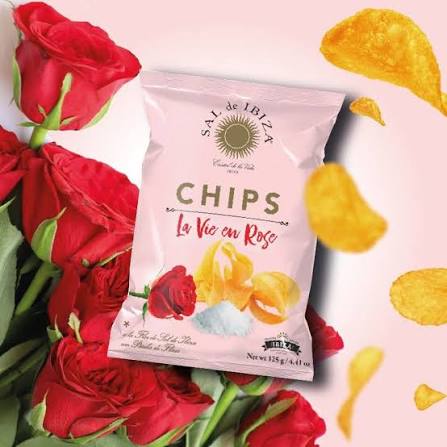A bag of Sal de Ibiza 'Chips La Vie en Rose' potato chips, styled with red roses and floating golden chips on a light pink background.