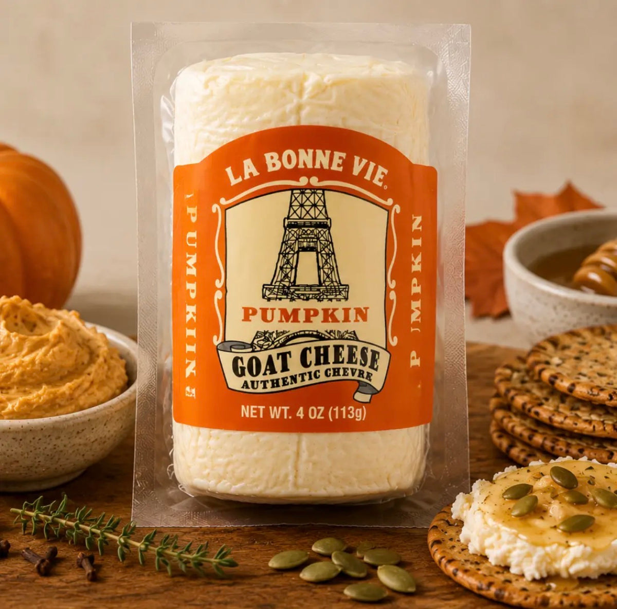 A 4 oz package of La Bonne Vie Pumpkin Goat Cheese stands on a wooden surface surrounded by autumn food props, including a bowl of pumpkin spread, cookies, a cracker with cheese and honey, pumpkin seeds, and a small pumpkin.