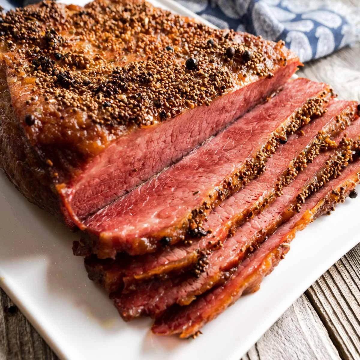 Sliced smoked brisket with a golden-brown seasoned crust and pink-red interior, served on a white square plate over a rustic wooden surface.