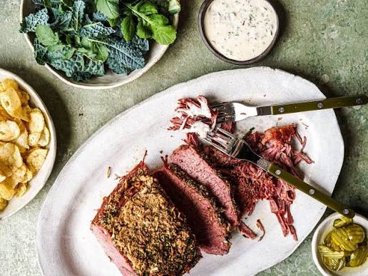 A plated slice of smoked corned beef with a bread crumb topping, served on a white oval platter with a fork, alongside side dishes of potato chips, kale, dipping sauce, and pickles on a green textured surface.