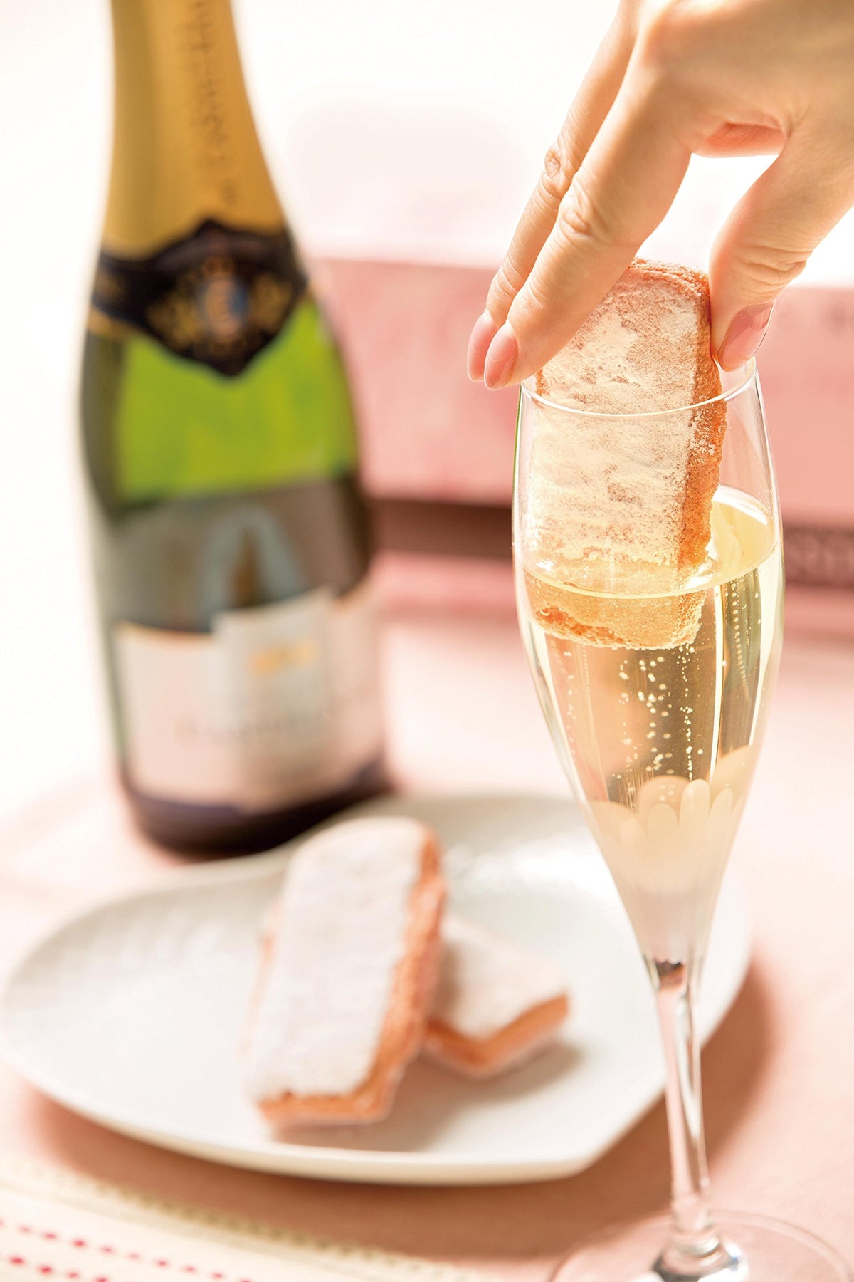 A hand holds a slice of pastry dipped in sparkling champagne over a filled flute glass, with a bottle and more pastries on a plate in the softly blurred background.