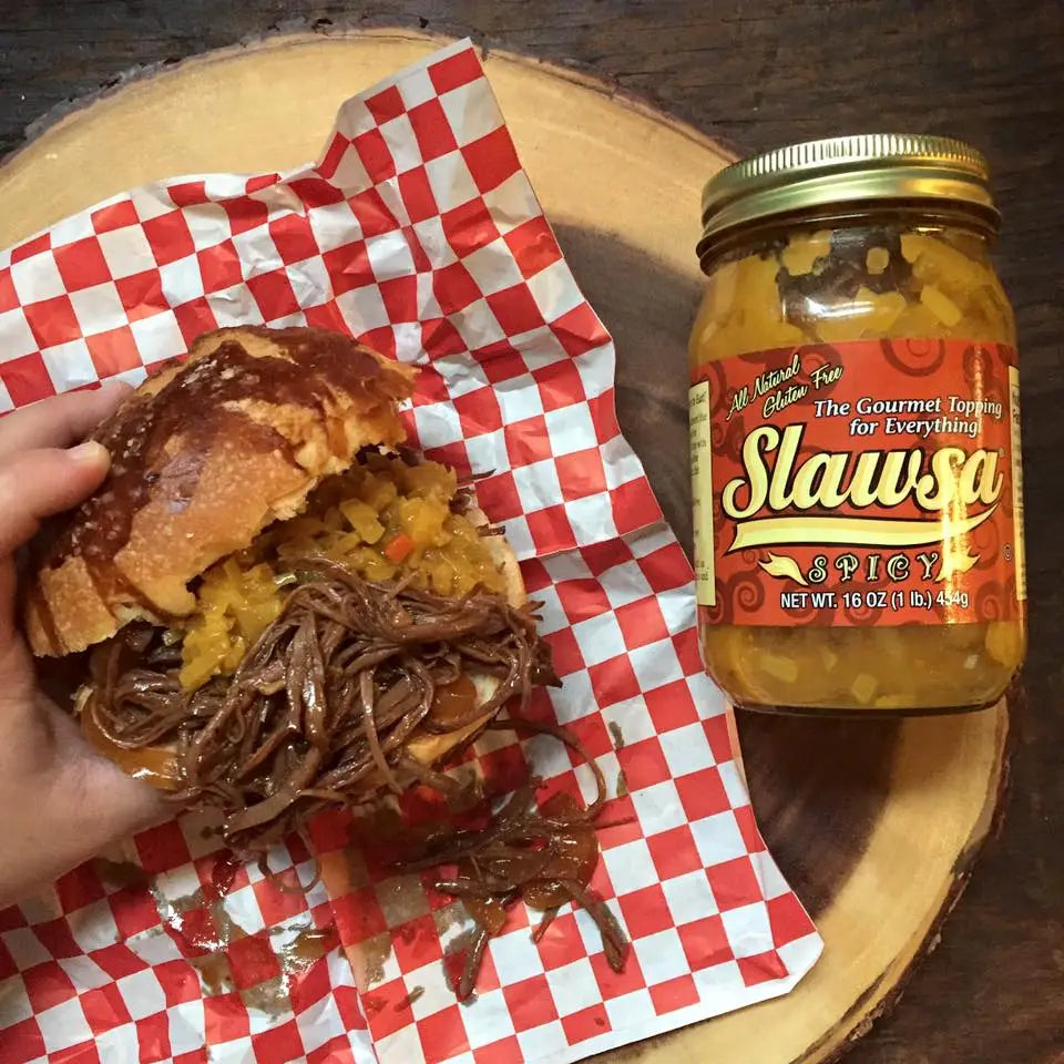 A hand holds a sandwich with pulled meat and yellow sauce next to a jar of Slawsa Spicy gourmet topping on a wooden board over a dark wood table.