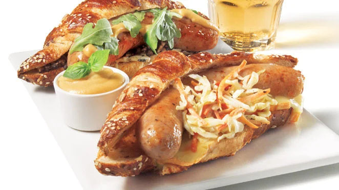 Two sausage-filled pretzel rolls with shredded cabbage, mustard and greens beside a glass of beer