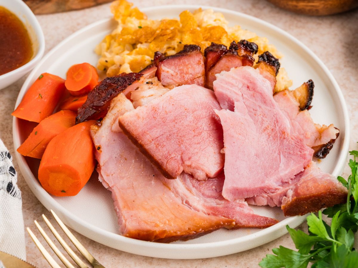 Sliced ham on a white plate with mashed potatoes, carrots, and a side of gravy, photographed in a real dining setting.