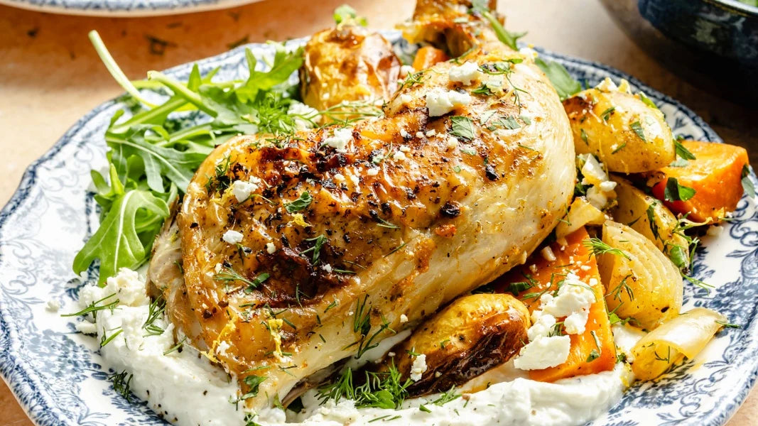 A close-up of a cooked chicken breast served on a blue-and-white patterned plate with arugula, roasted potatoes, and a white creamy sauce.