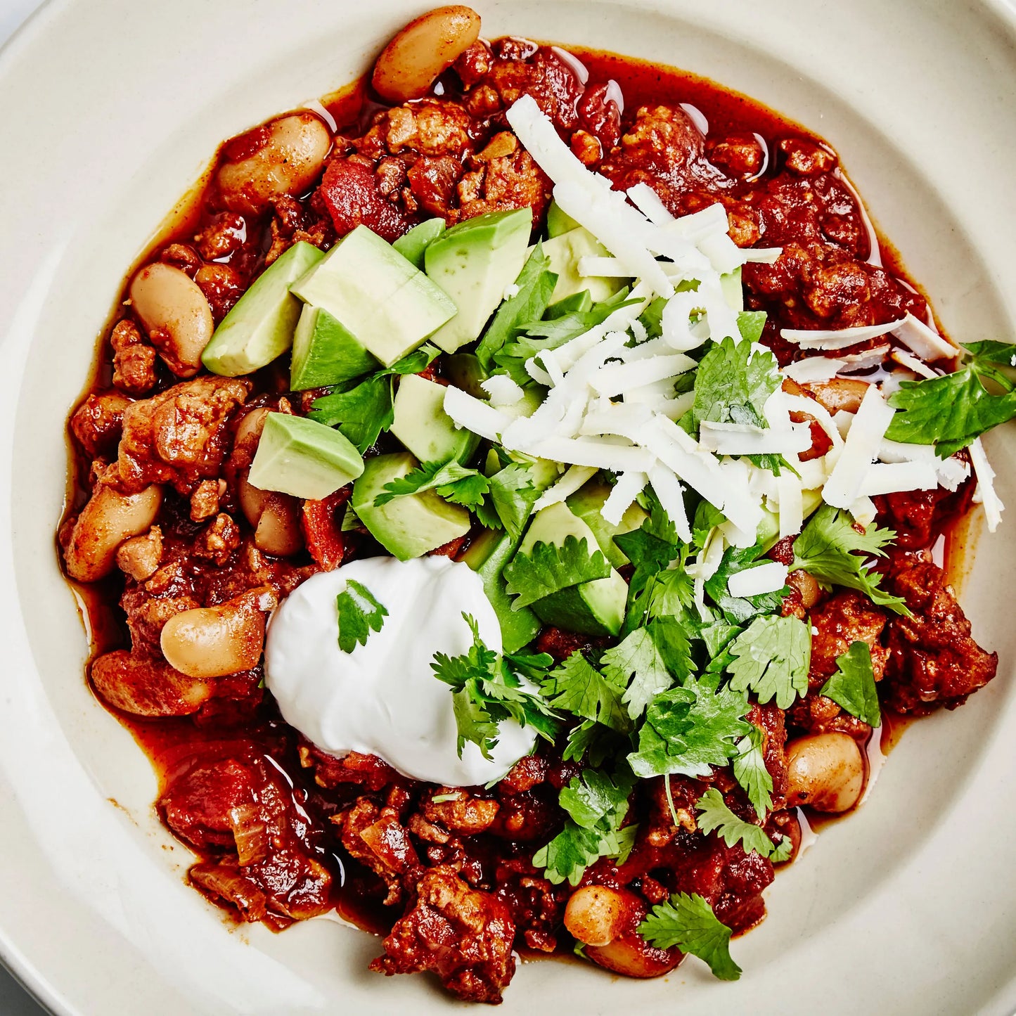 A close-up overhead shot of a bowl of chili con carne ground chicken chorizo  topped with diced avocado, shredded white cheese, a dollop of sour cream, and fresh cilantro, served in a light beige bowl.