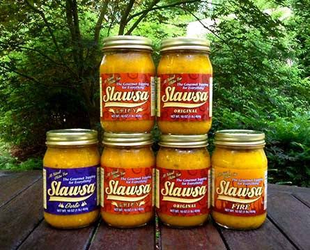 Six jars of Slawsa hot sauce arranged on a wooden table outdoors, with green foliage in the background. The jars contain orange-red sauce and have gold lids, with labels in red, blue, and white.