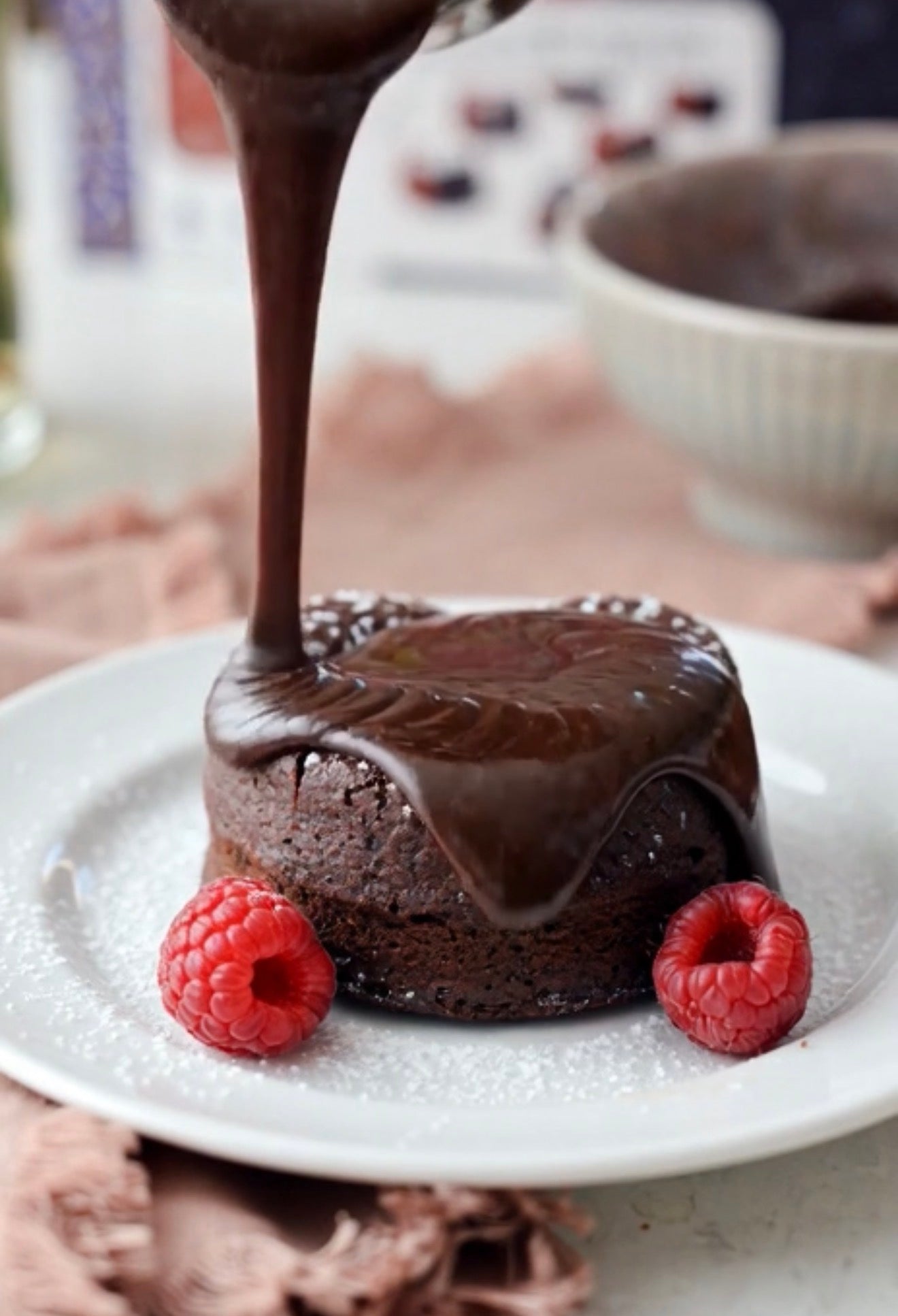 A chocolate lava cake on a white plate, with thick chocolate sauce being poured over it from above, garnished with two fresh raspberries and a dusting of powdered sugar.