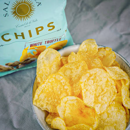 A bowl of golden yellow potato chips with white truffle seasoning, shown next to a light blue bag of Salta Chips on a gray fabric background.