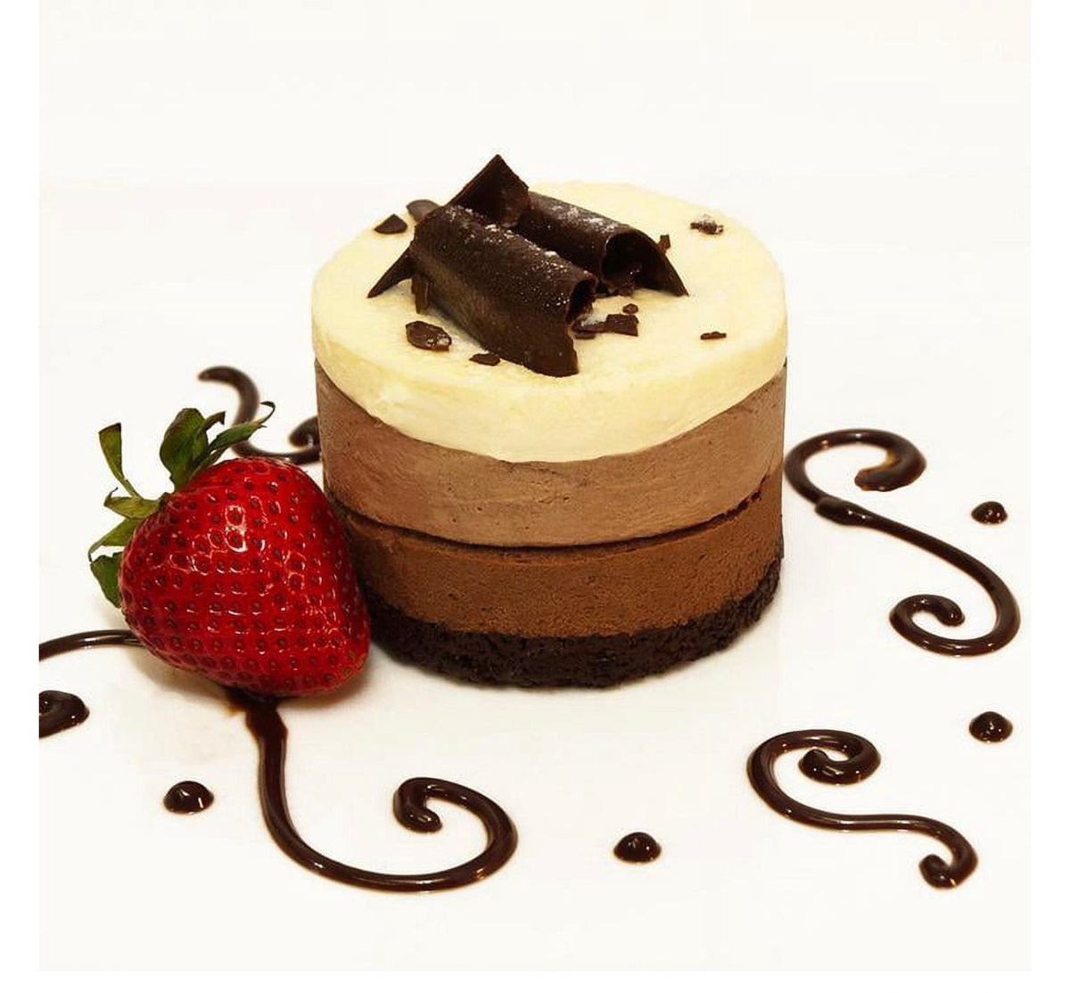 A small three-layer chocolate mousse cake with a dark chocolate base, cream-colored top layer, chocolate curls on top, and a fresh strawberry beside it on a white background with chocolate sauce drizzle.