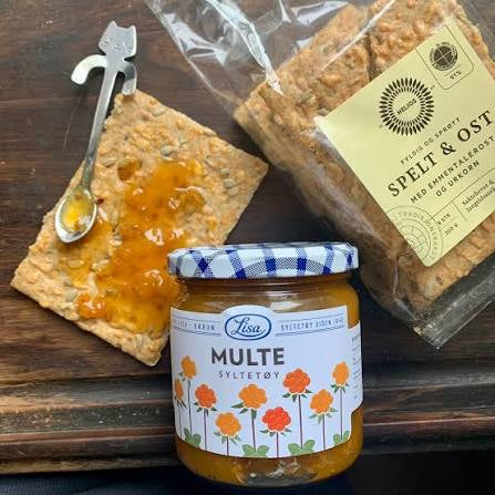 A jar of Lisa Multe Syltøy orange spread in the foreground, with a graham cracker topped with orange spread and a spoon on the left, and a package of Sara Spelt & Oost herb enriched wheat flour on the right, all arranged on a dark wooden surface.
