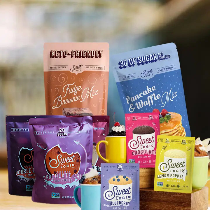 A collection of Sweet Logic keto-friendly coffee and dessert mixes displayed on a wooden shelf with colorful mugs. The products include Fudge Brownie Mix, Pancake & Waffle Mix, Chocolate Chip Cookie Mix, Blueberry Pancake Mix, Lemon Paddy Mix, and Double Chocolate Cookie Mix, shown in various vibrant packaging colors like blue, purple, pink, and yellow.