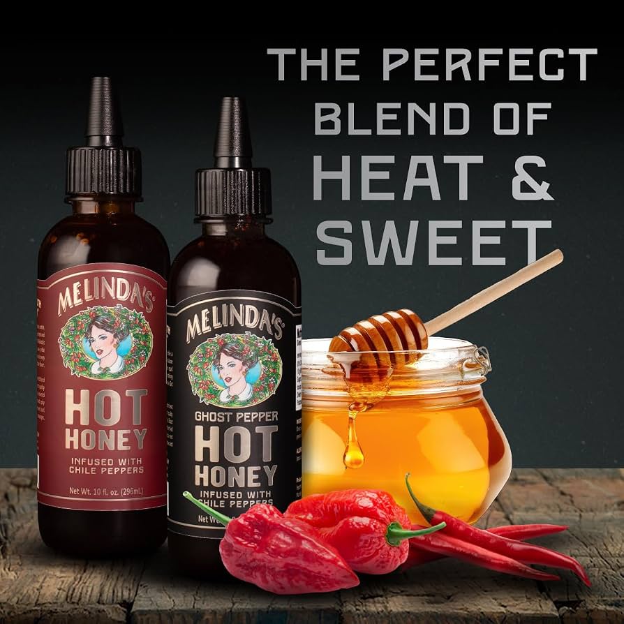 Melinda’s Classic and Ghost Pepper Hot Honey bottles beside a honey jar, honey dipper, and chile peppers
