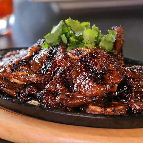 Grilled short ribs with a glossy dark sauce, served on a black cast-iron plate with fresh green cilantro garnish, set on a wooden surface.