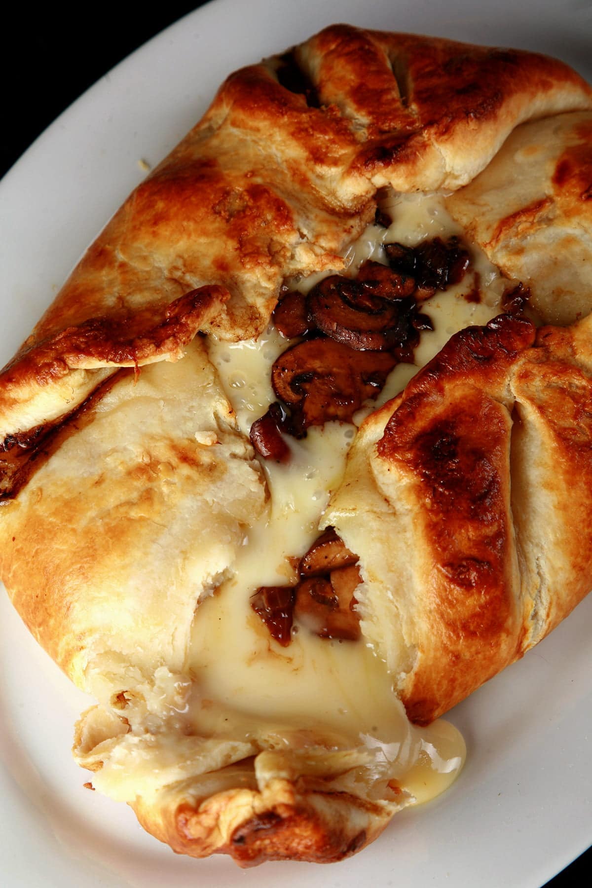 A close-up of a baked pastry or bread roll on a white plate, with golden-brown crust, melted cheese, and browned mushroom pieces visible inside.