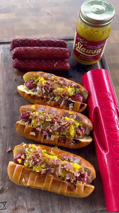 Three hot dogs topped with mustard, onions, and bacon are arranged on a wooden board next to three grilled sausages and a jar of Slawsa relish. A bright pink silicone lid is also visible on the right side.