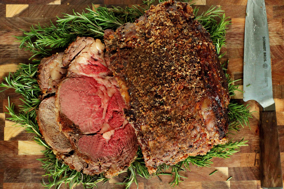 A sliced roast of cooked beef prime rib roast  and browned exterior, arranged on a wooden cutting board with green rosemary sprigs and a carving knife.