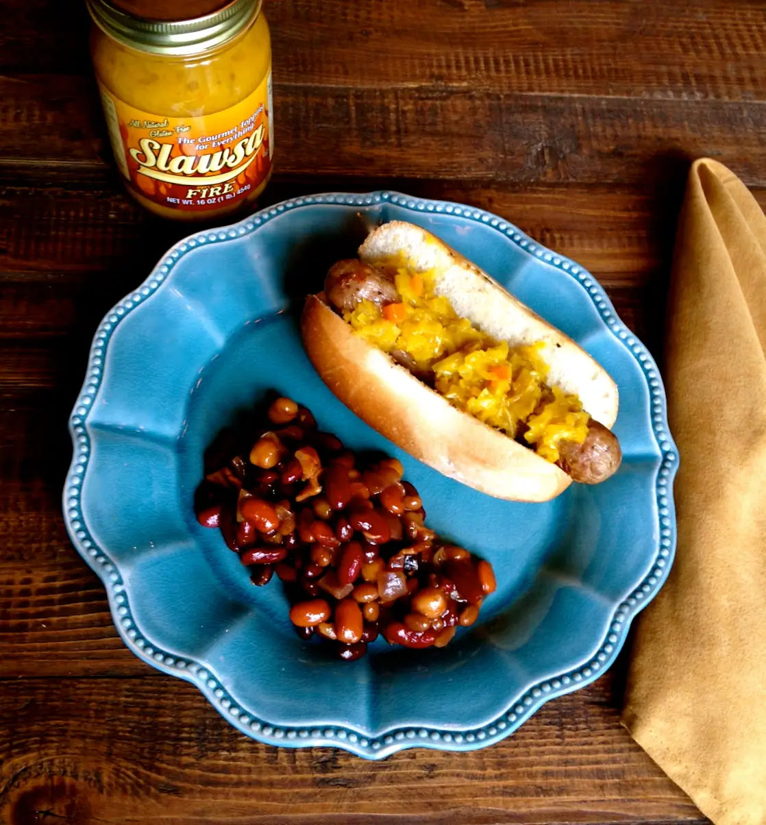 A blue scalloped plate holding a hot dog topped with mustard and a side of baked beans, set on a wooden table with a jar of Slawsa Fire mustard and a tan napkin nearby.