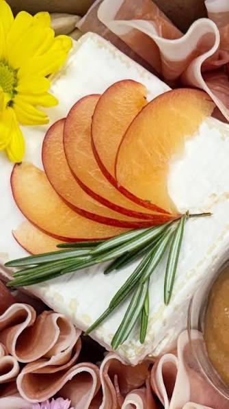 A close-up styled food scene showing a rectangular block of Trillium cheese topped with sliced peach or apricot wedges, a green rosemary sprig, and rolled pink cured meat around it, with a yellow flower in the corner.