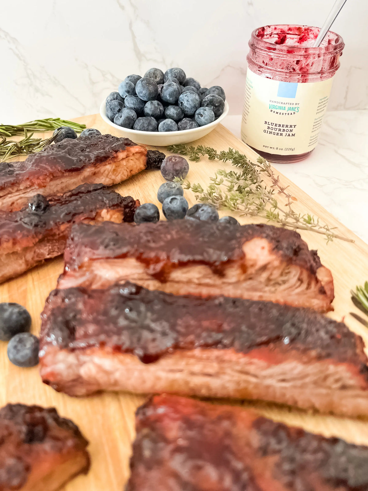 Ribs glazed with dark berry jam on a wooden cutting board, with a bowl of blueberries, a jar of blueberry raspberry ginger jam, and herbs in a kitchen setting.