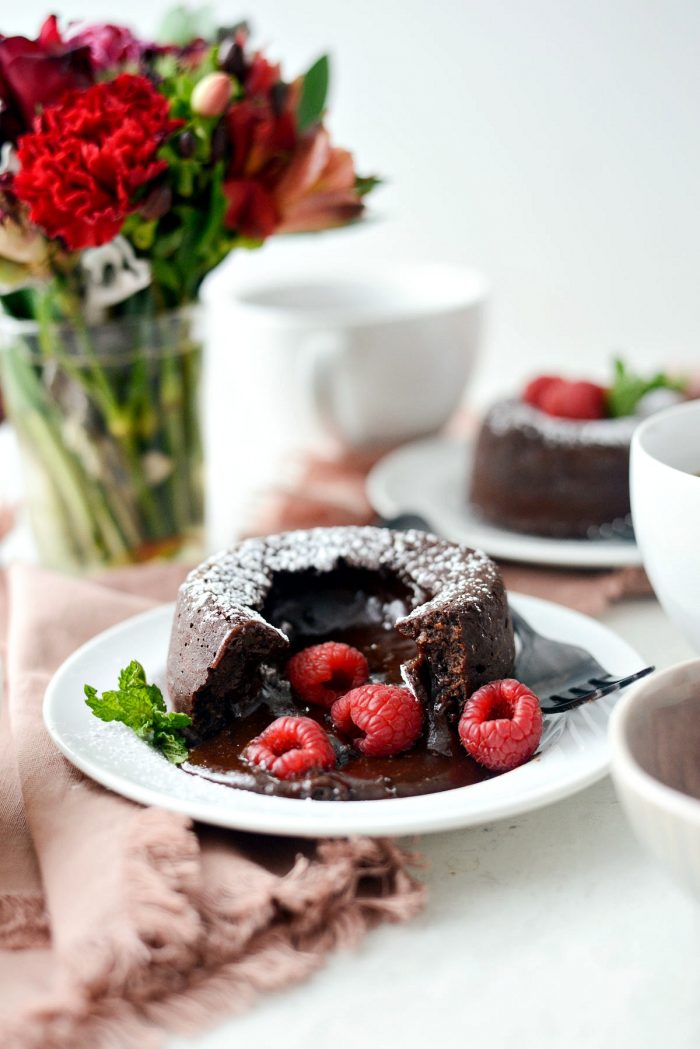 A chocolate lava cake on a white plate, topped with fresh raspberries and a mint leaf, with a blurred background showing more cakes, cups, and a vase of flowers.
