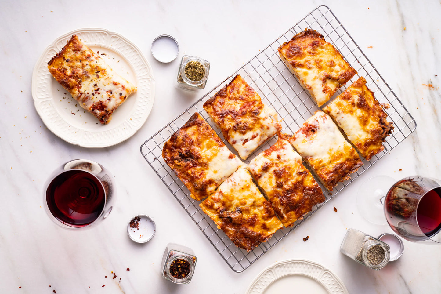 A top-down view of baked rectangular cheese-topped bread or flatbread pieces arranged on a wire cooling rack, with a few slices on plates and small condiment containers on a light marble surface. No person or animal is present, but this is not a clean isolated studio product shot because it includes styled food props and a real tabletop setting, so Lifestyle fits better than Product/Packaging.