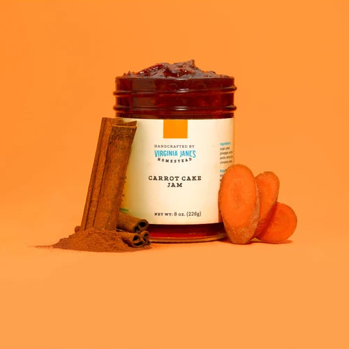 A jar of Virginia Jane's Remembers Carrot Cake Jam on a solid orange background, styled with cinnamon sticks, ground cinnamon, and sliced carrots.