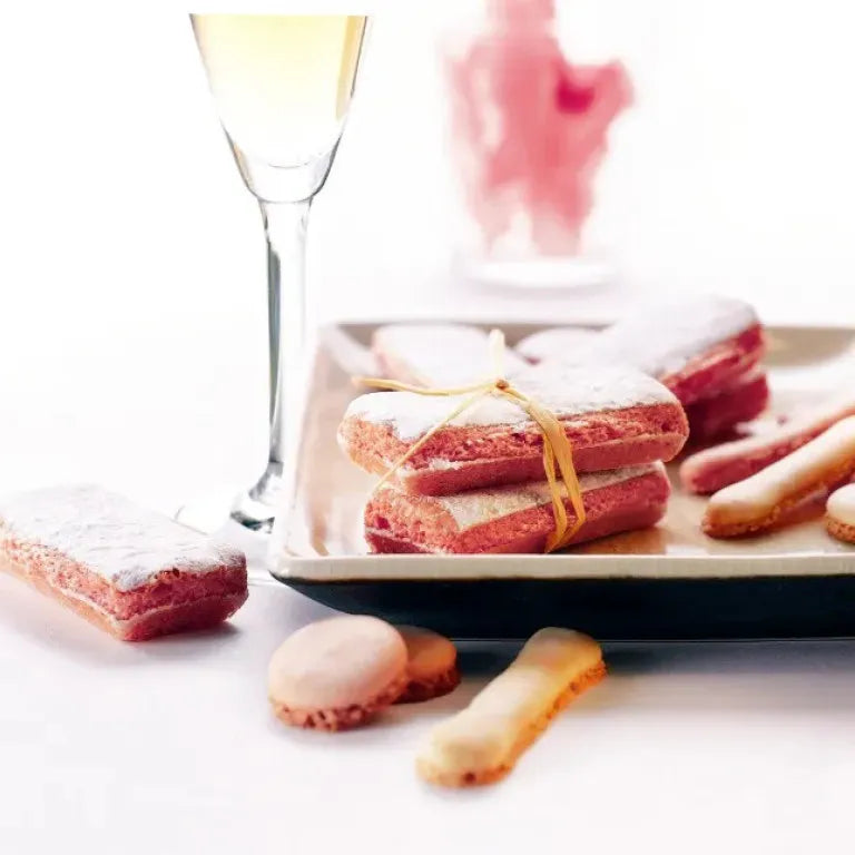 Pink sponge cookies with white cream filling, dusted with powdered sugar and tied with a yellow ribbon, arranged on a rectangular plate with ladyfinger biscuits and macarons on a white surface, next to a glass of white wine.