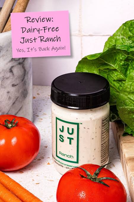 A jar of Just Ranch dairy-free dressing with a black lid and cream-colored contents, styled on a kitchen counter with tomatoes, carrots, lettuce, and a pink review note.