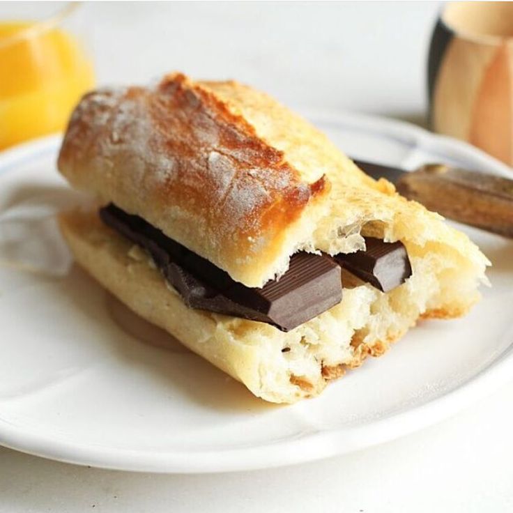 A close-up of a baked pastry sandwich filled with chocolate bars, served on a white plate with a blurred breakfast background.