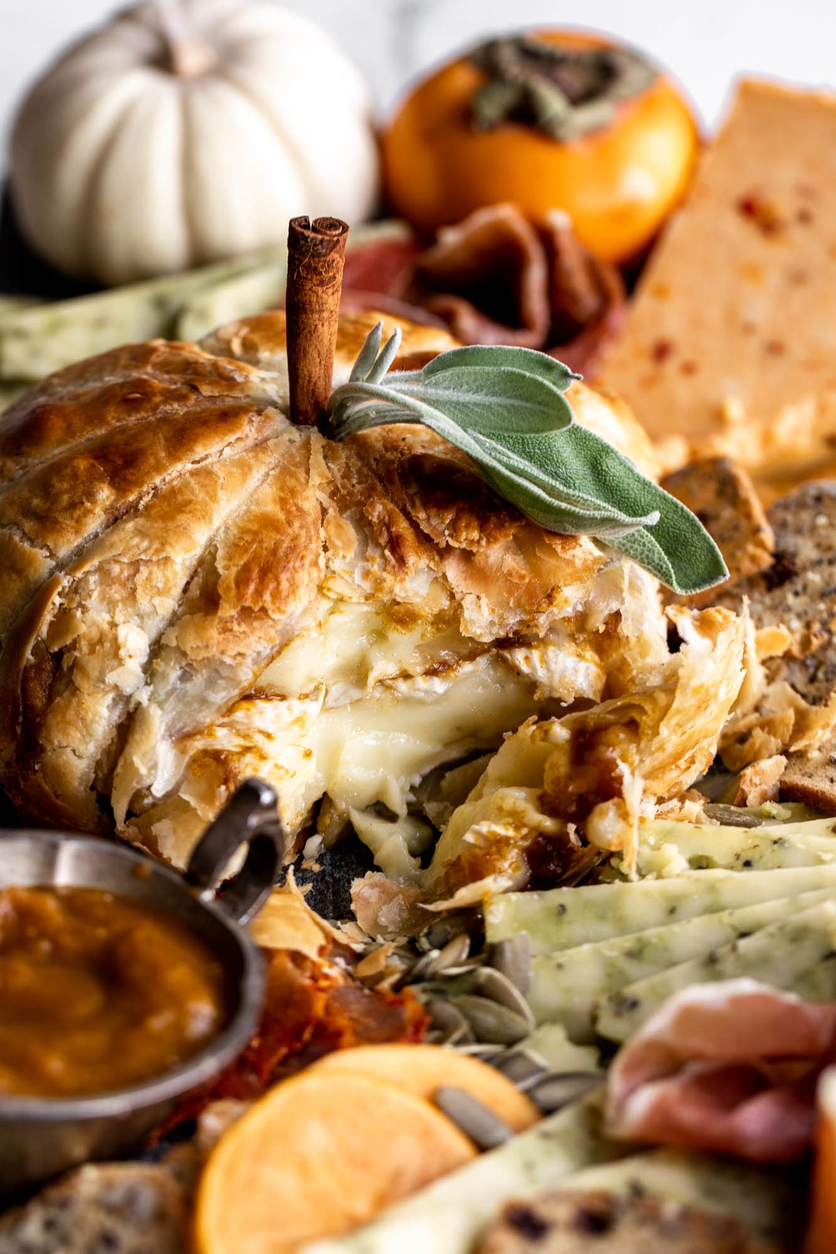 A close-up food scene featuring a baked pumpkin bread or cheese ball with a cinnamon stick and sage garnish, surrounded by cheeses, crackers, persimmon, and a small bowl of sauce on a light background.